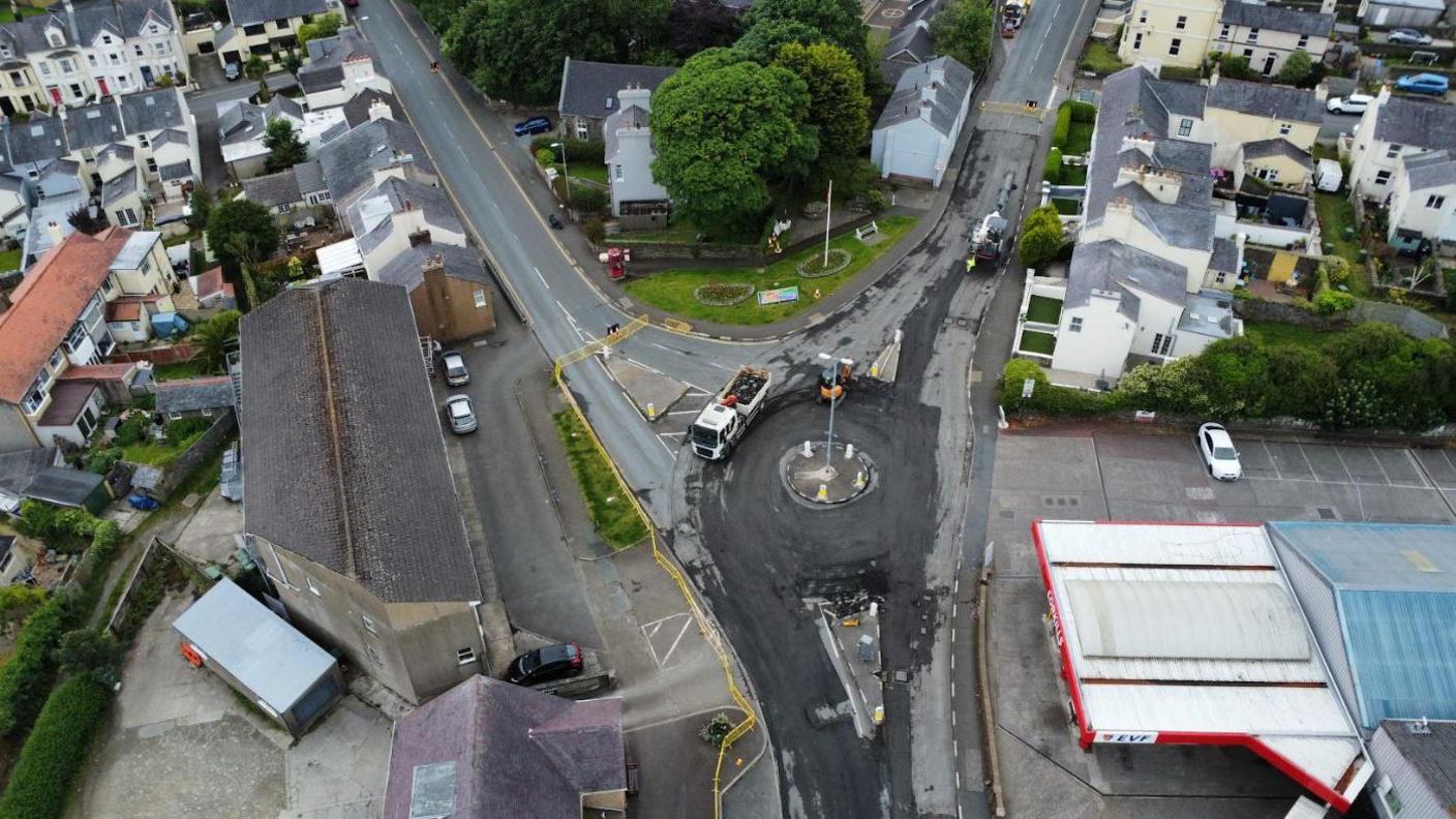 An aerial view of resurfacing works at Corkills Roundabout. Three roads can be see joining the roundabout, with the junction flanked by a petrol station, large houses and a big commercial building, which is set back from the road with a parking area in front of it. 