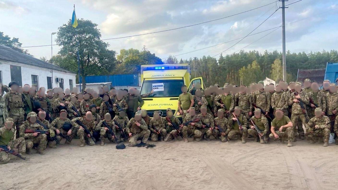 A lime green ambulance at a military base in Ukraine with about 60 soldiers in army uniform. Their faces are obscured for security reasons.