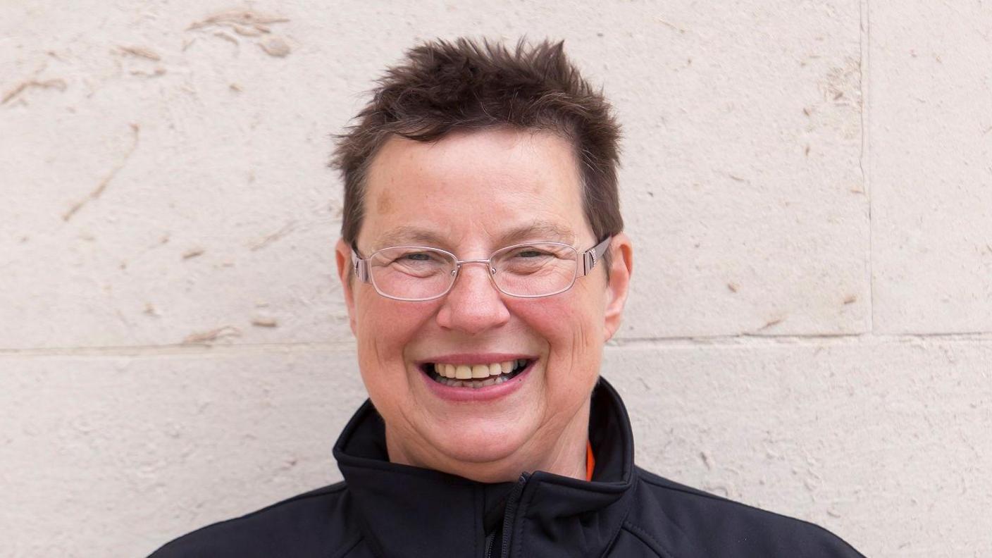 A woman with short brown hair and glasses smiles at the camera. She is standing in front of a white wall and wearing a black jacket.