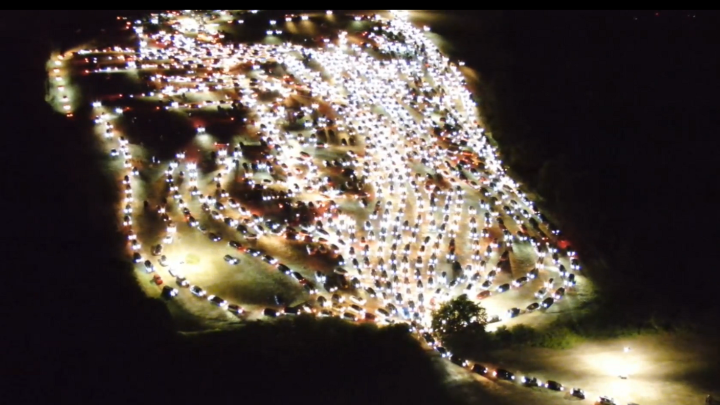 Hundreds of cars queuing to exit a venue at night. It is pitch black. Their headlights are on. There is only one exit.
