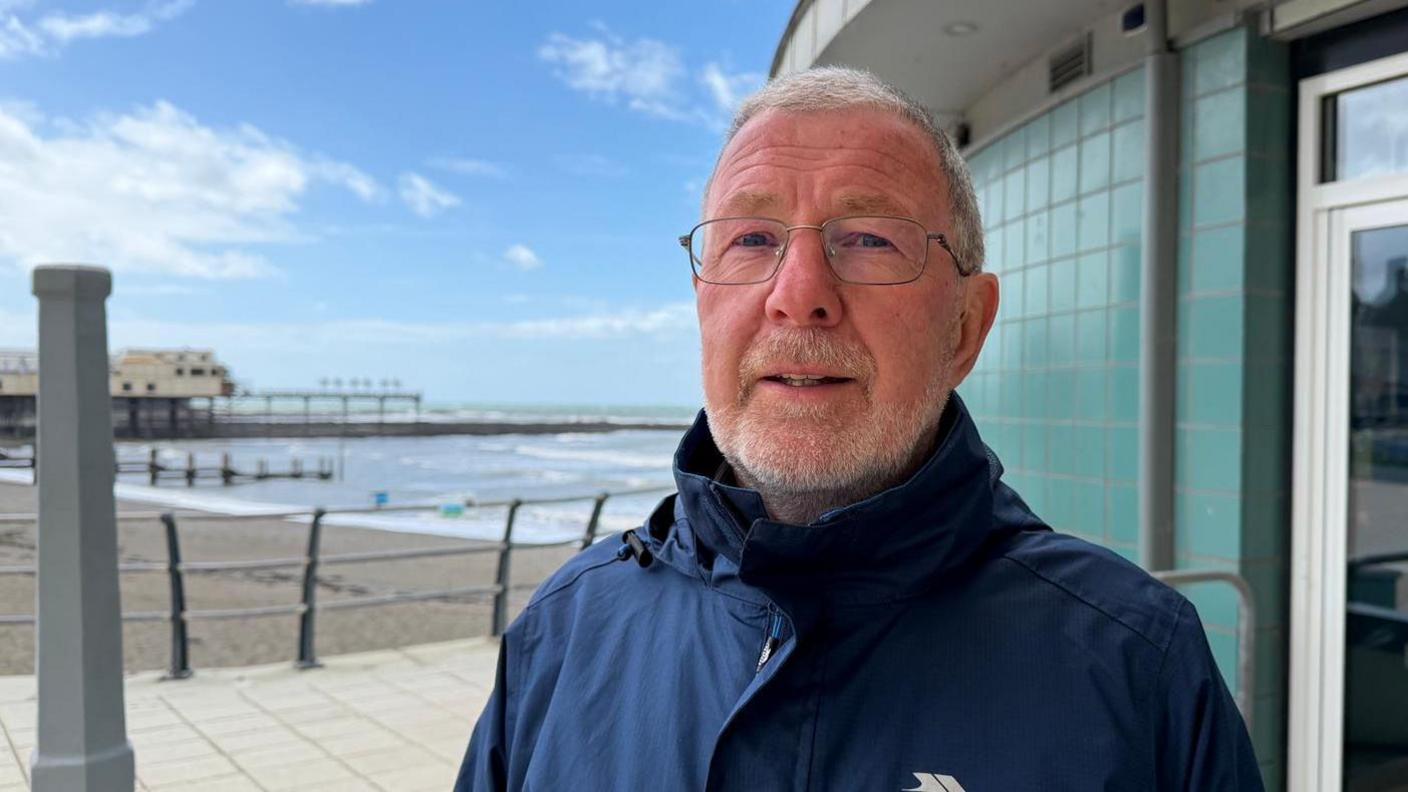 Llun o'r cynghorydd sir Gareth Davies yn sefyll ar bromenad Aberystwyth - dyn a barf, sbectol yn gwisgo cot las, y mor a pier Aberystwyth yn y cefndir