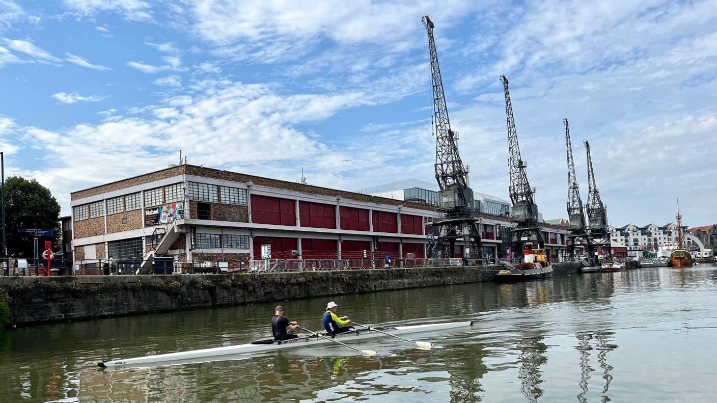 Two men row across Bristol Harbour in a slender rowing boat. Behind them the cranes of the Mshed Museum are visible. The sun is largely blue, with patchy white cloud