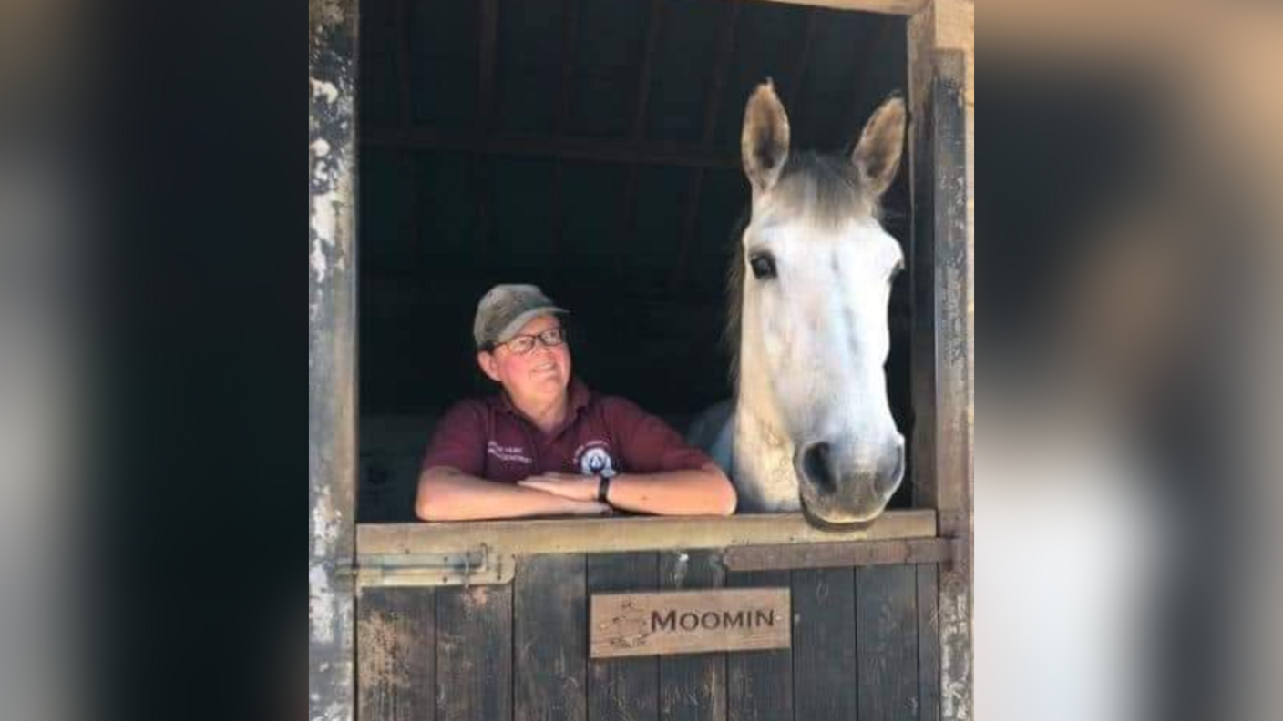 Jayne Hunt standing next to a white horse. She is wearing glasses, a hat and a burgundy polo shirt. Her arms are on top of a stable door and she is looking at the horse.