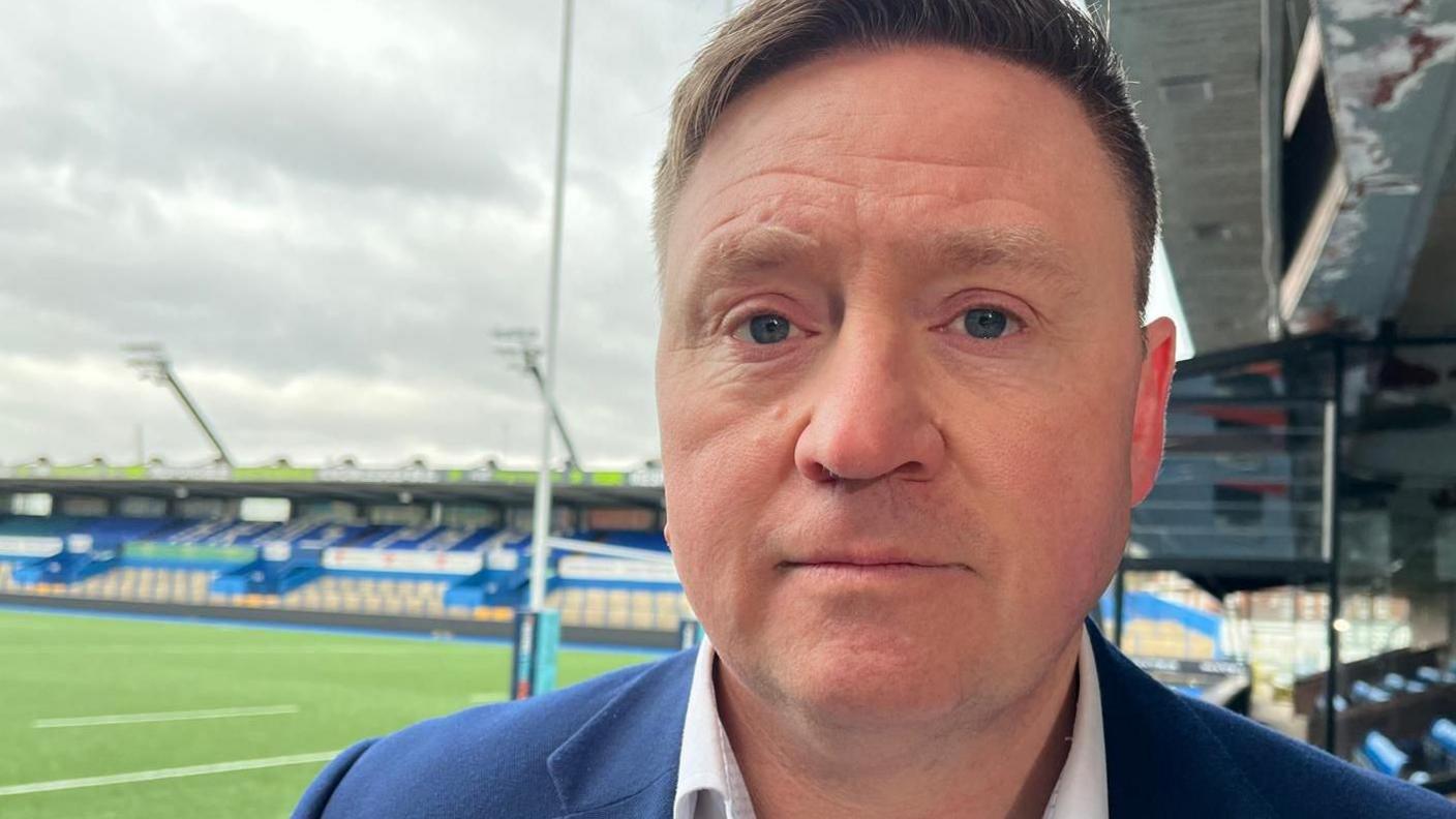 Rhodri, who has short dark hair, is wearing a navy blazer and white shirt. In the background is the empty stadium and pitch of Cardiff Arms Park. 