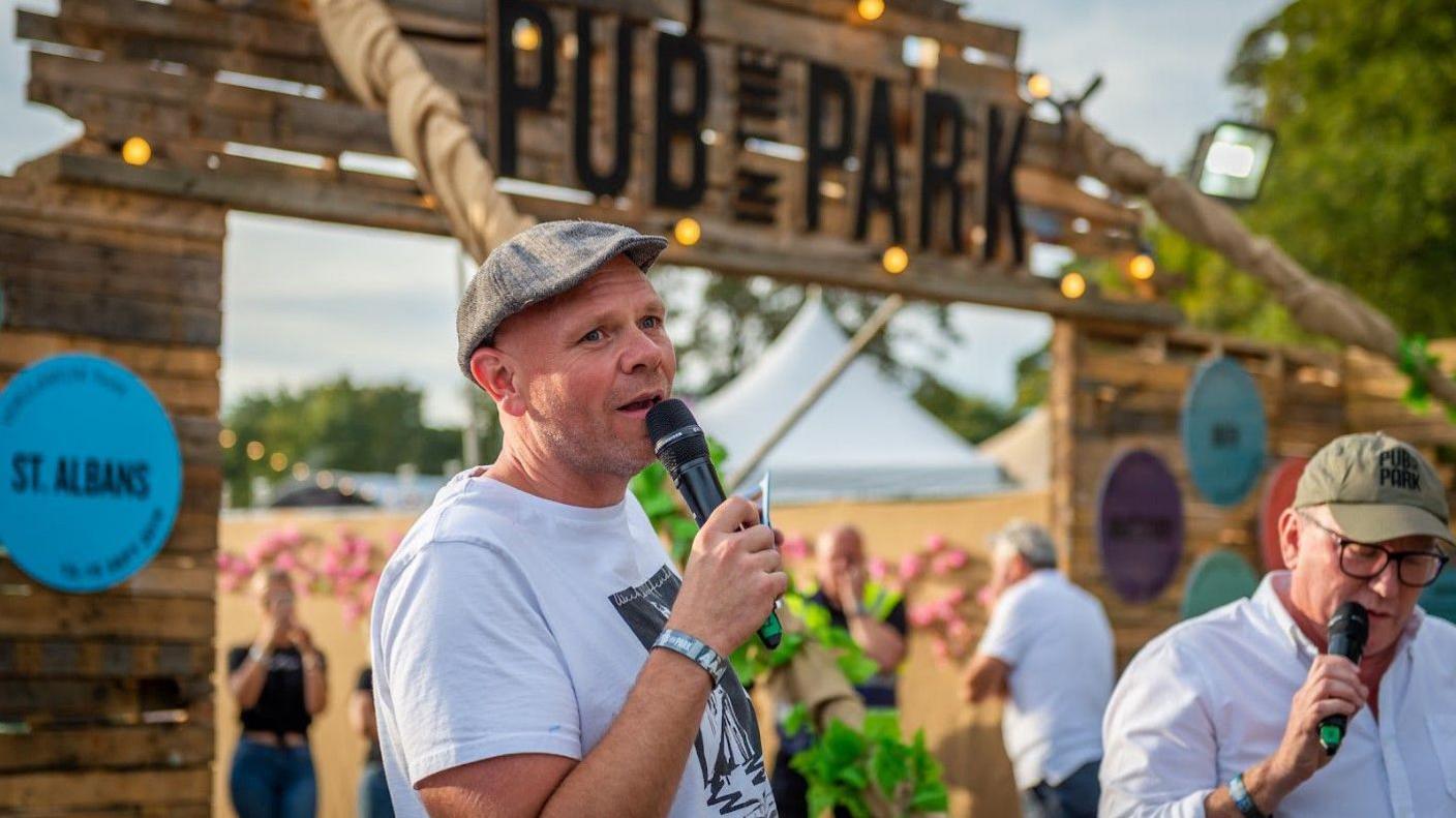 Chef Tom Kerridge wearing a light grey T-shirt and darker grey flat cap, holding a microphone in front of a sign saying Pub in the Park in St Albans