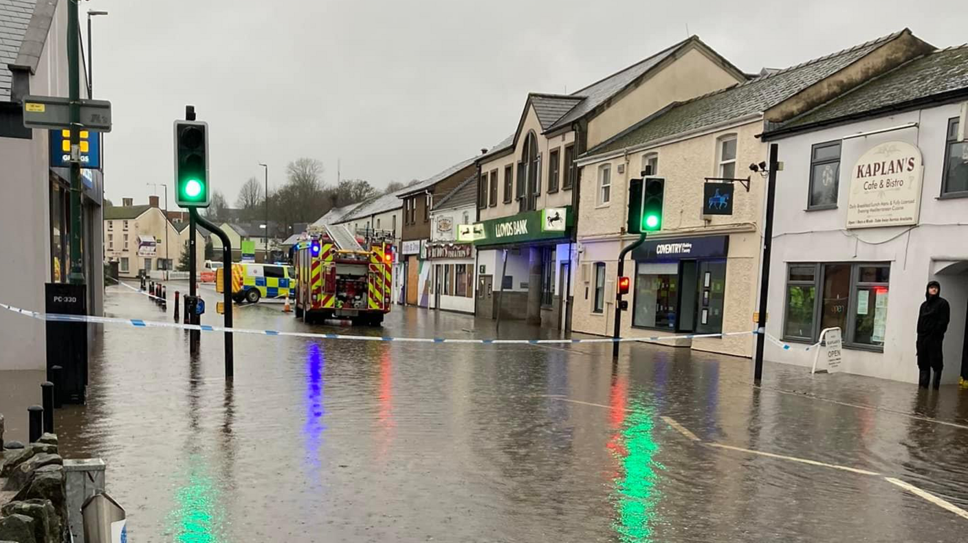 A street through Lydney in Gloucestershire is seen flooded with a fire engine visible in the distance. The water is several inches deep