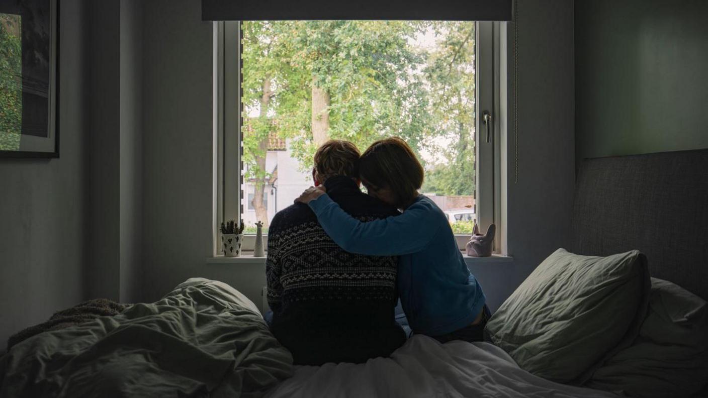 A posed picture of two adults, a man and a woman, sitting on a bed at a window. The woman is hugging the man as he looks out the window