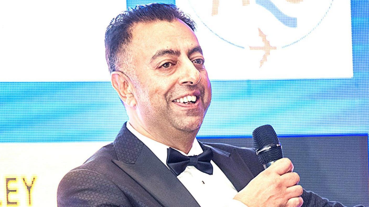 A man in his fifties, in a black suit and tie and white shirt. There is what appears to be a large LED screen behind him and he is holding a microphone, seemingly on stage. He is smiling. He has black hair, short on the sides and pushed back on top.