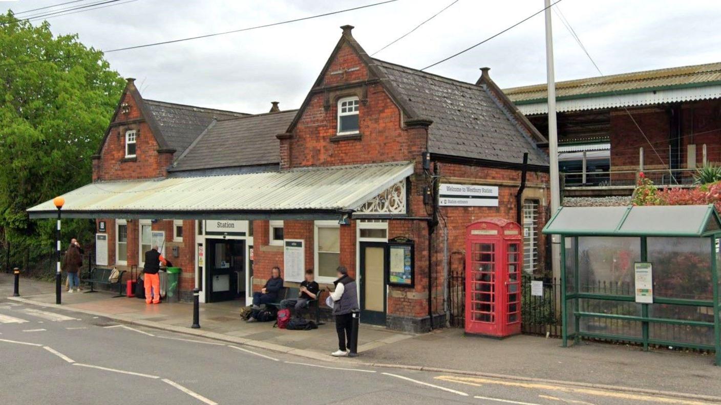 The entrance to Westbury train station. It is a small red brick building with a red phone box outside. There is a clear plastic veranda on the front exterior where people are stood waiting. You can see the railway line in the background.