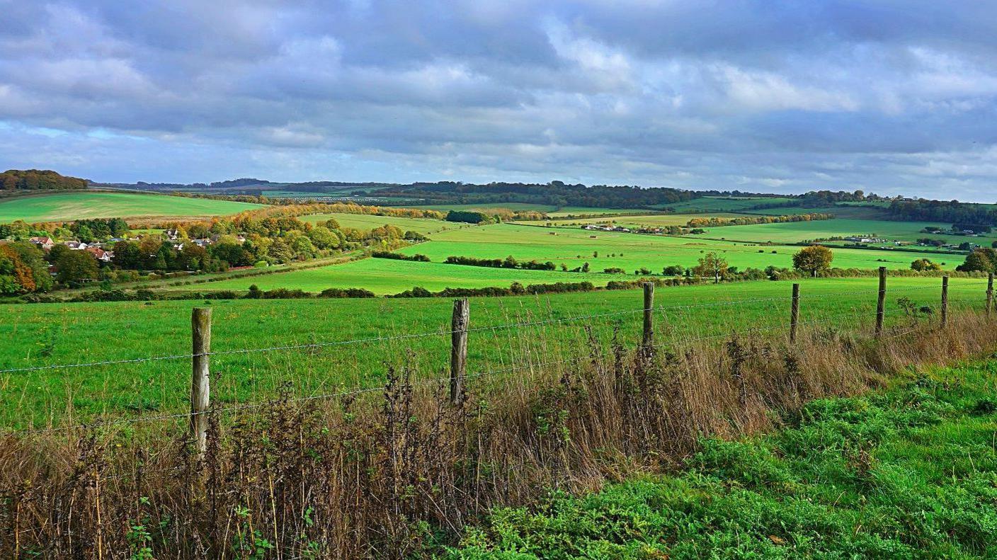 View of a fence between two fields and a wide view of green rolling hills with some hedges under a sunny sky with some cloud.