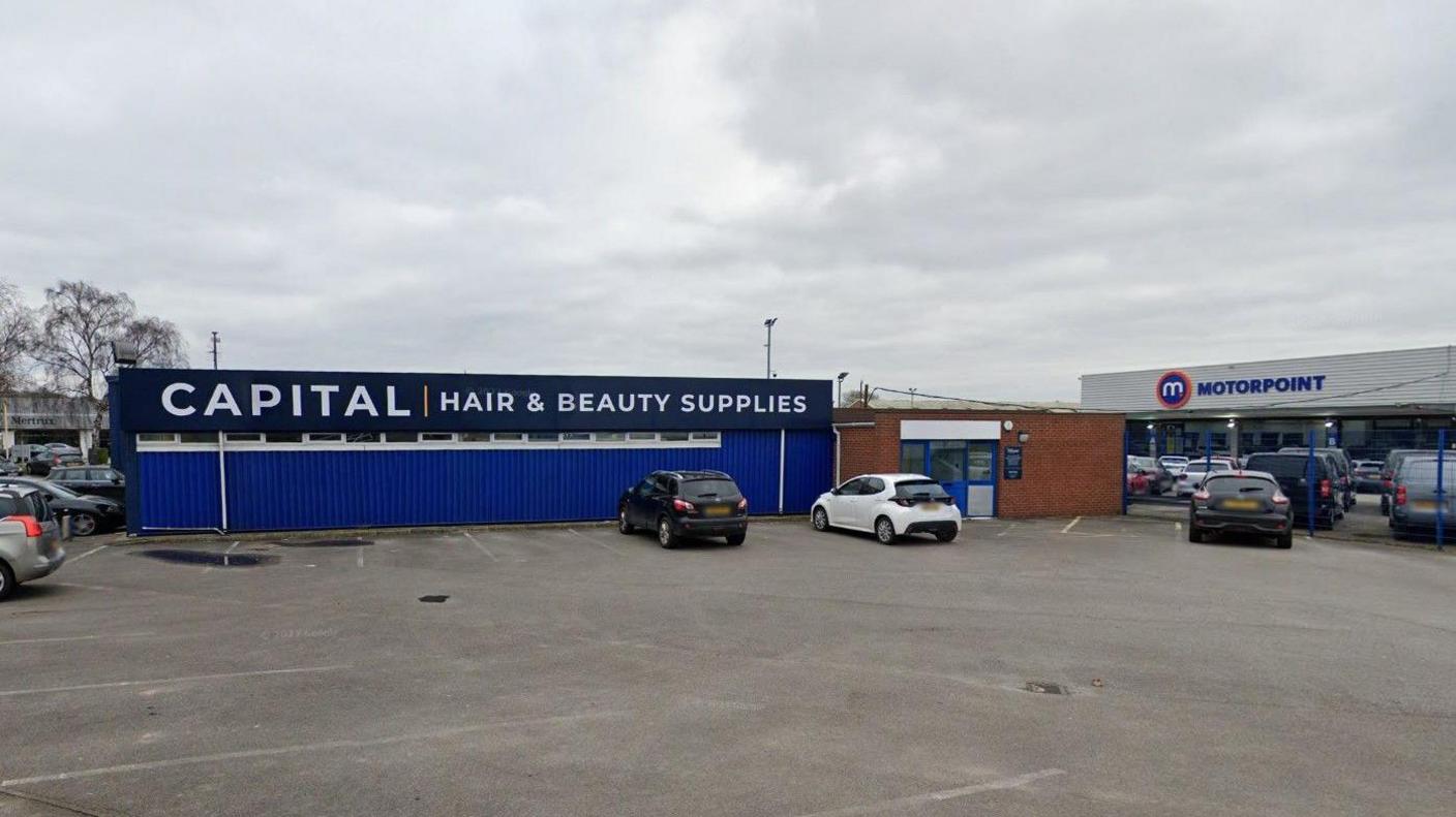 A street view of hair and beauty supplier business. The one-storey building is bright blue warehouse with a sign that reads 'Capital, hair and beauty supplies'. Two cars are parked directly in front of the building. A car sales warehouse can be seen in the background.