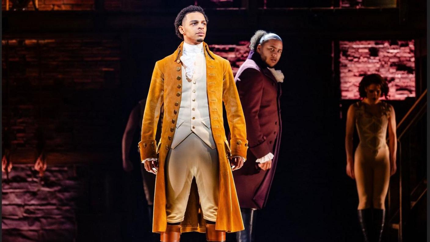 Marley Fenton, 23, on stage and in costume as Alexander Hamilton in a long yellow velvet coat and white waistcoat