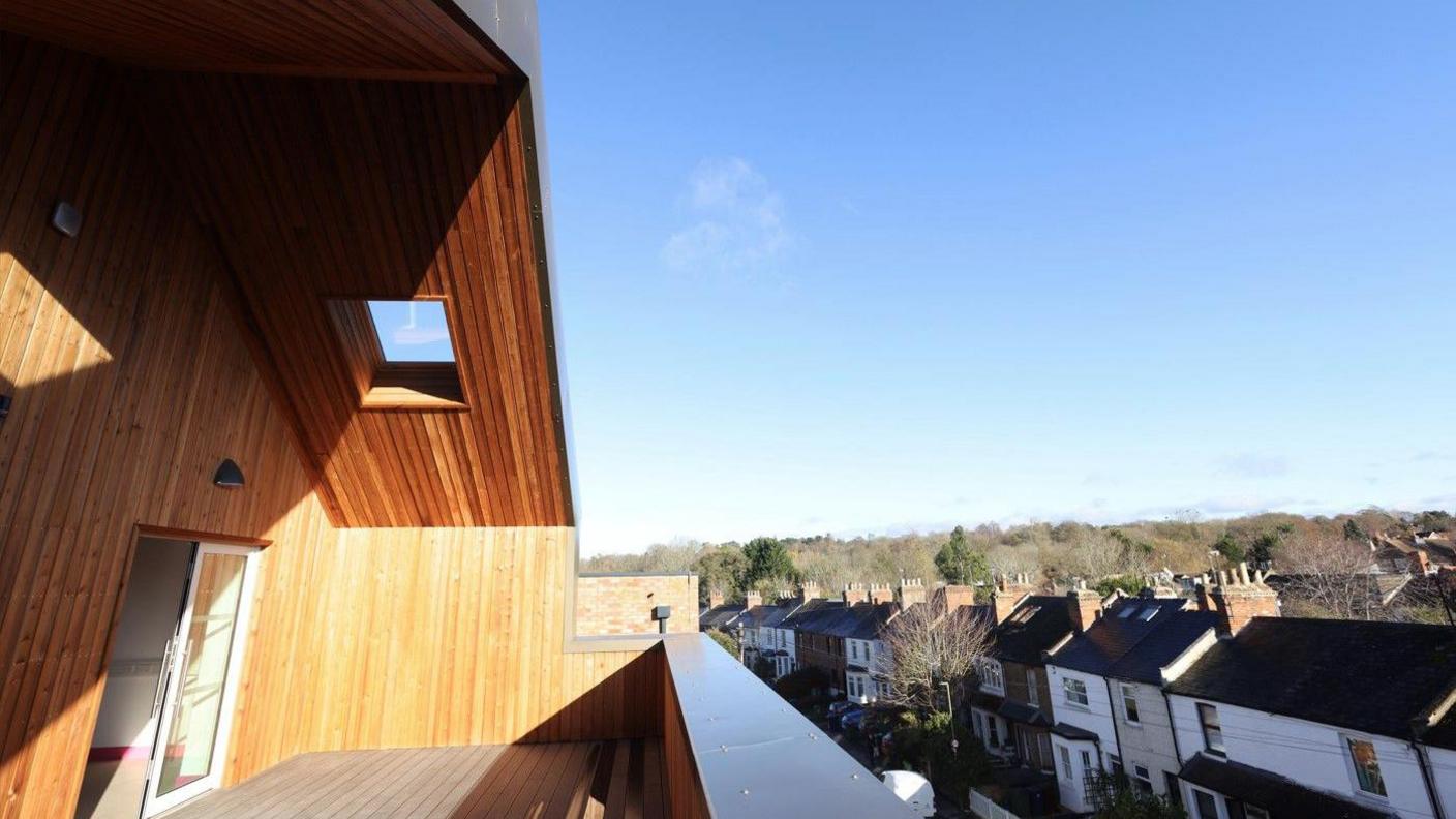Under a bright blue sky, a third floor wood-lined balcony overloooks rows of terraced houses.