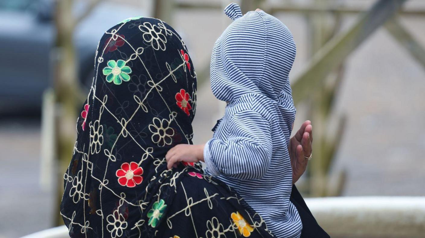 A woman wearing a head scarf with flowers on it holds a young child who has a blue and white stripey hooded jumper on.