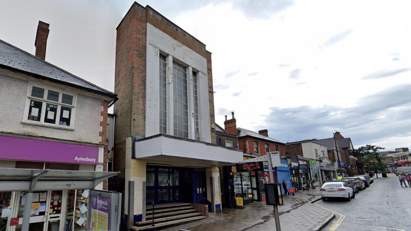 Aylesbury High Street, showing a former bingo hall, rows of shops and a street with cars parked on it 