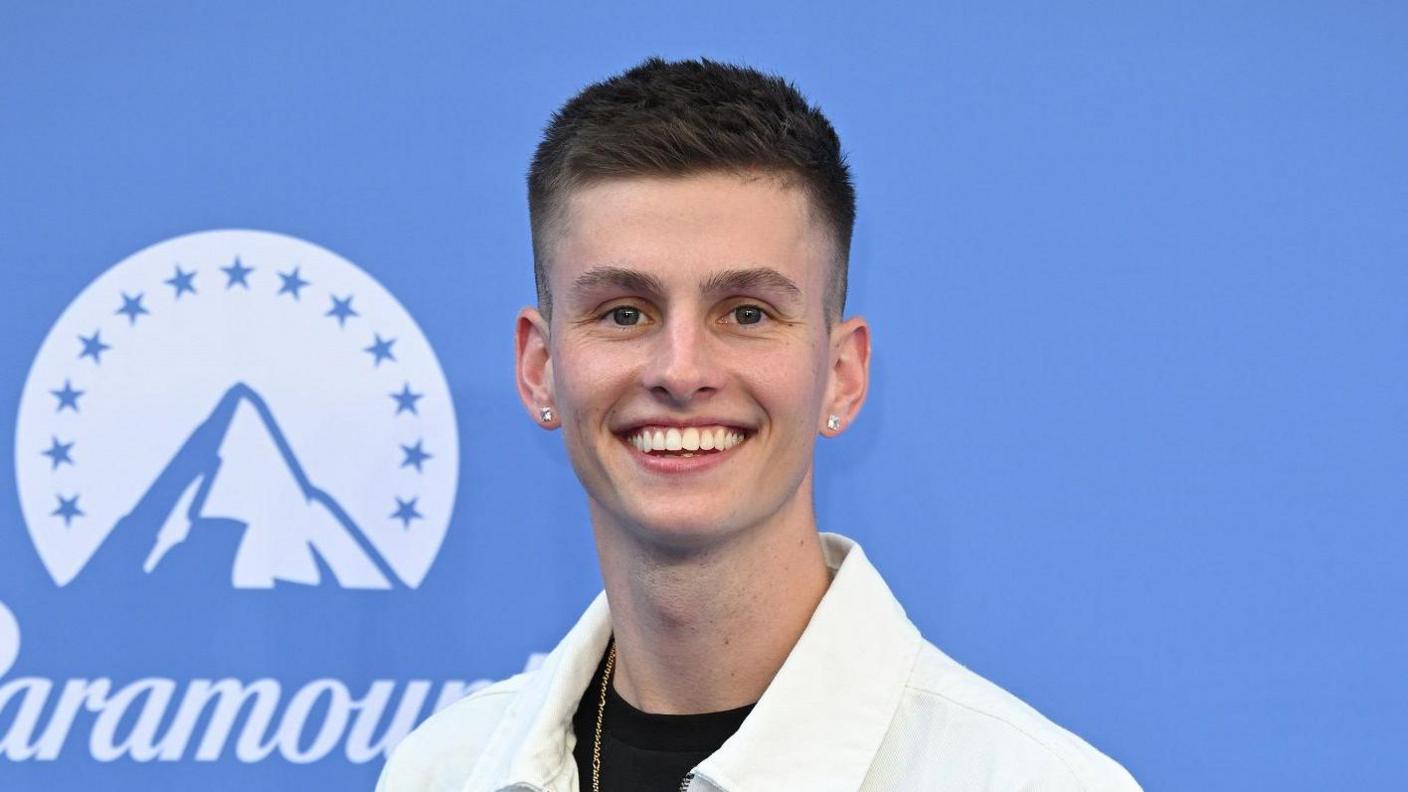 A man with short brown hair wearing a white shirt stands in front of a blue background with a Paramount logo on it
