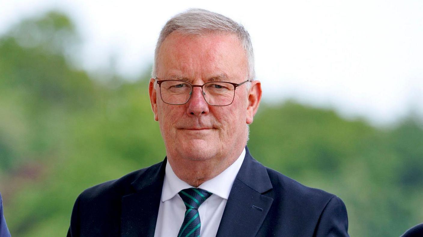A man with grey hair wearing thin-framed black glasses.
He is wearing a navy jacket, white shirt and a navy and green diagonal striped tie.