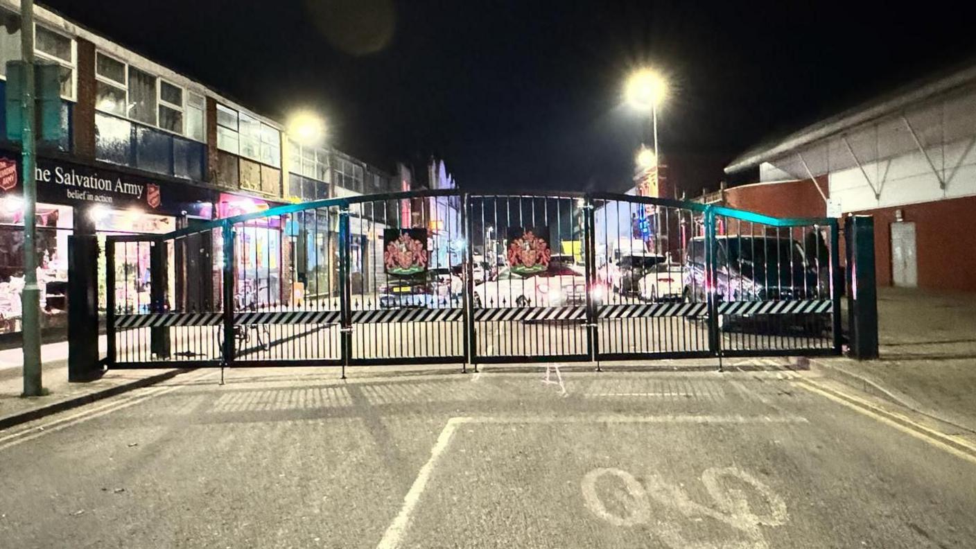 Closed bi-folding gates on Eastgate Street. They are black with the city's coat of arms - two red lions and a red shield - on each of the two bi-fold gates. 
Behind the gates is a road with cars parked either side of it. 
The picture was taken when they were closed so is during the late evening and it is dark outside.
There are shop fronts on each side of the road, with The Salvation Army clearly signed on the left of the picture.