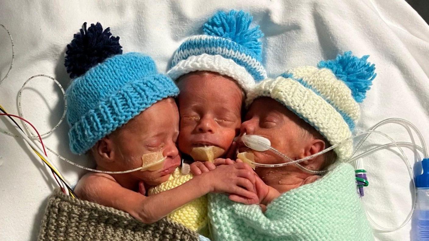 The trio lying next to each other holding hands in the hospital unit. They are all wearing blue hats and have medical tubes attached, with one baby wrapped in a brown blanket, one in yellow and one in green