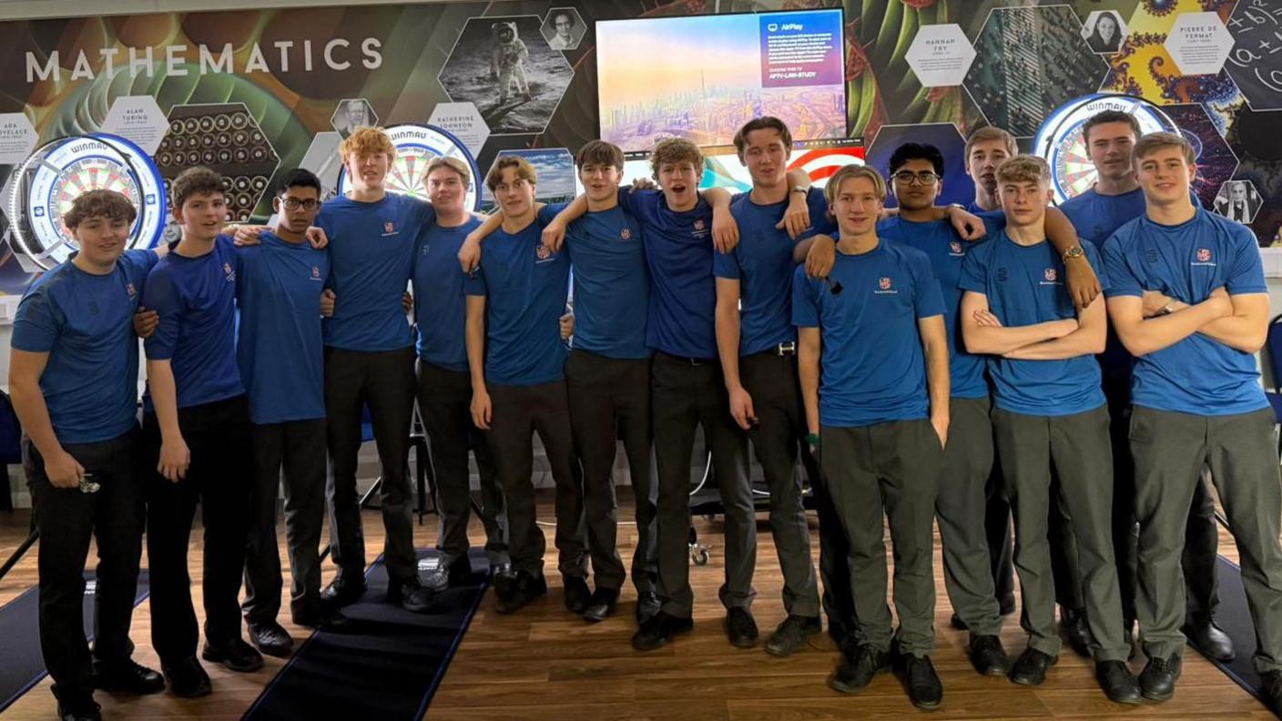 A group photo of 15 boys, all in blue school-branded T shirts with their arms around each other