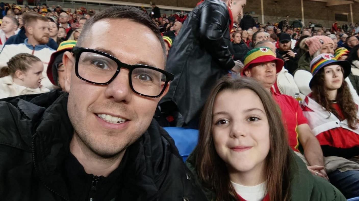 Steve and Eliza are in the crowd at a football match. Steve, left, has a black coat and is wearing glasses. Eliza, right has long brown hair and a green coat over a Wales top. 