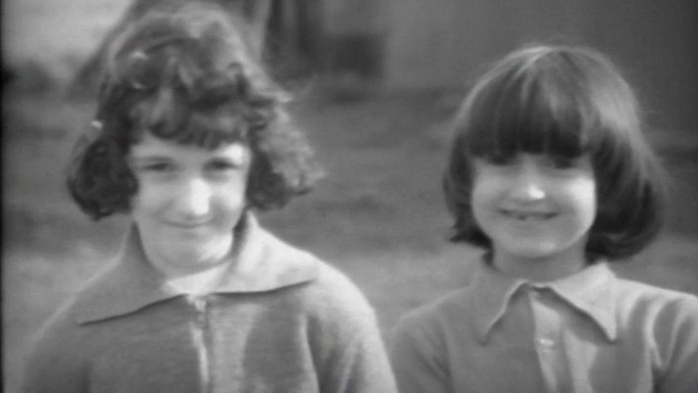 Two young girls smile at the camera, in black and white. They have brown hair cut in short styles and are wearing wool shirts. The one on the right has a tooth missing at the front.