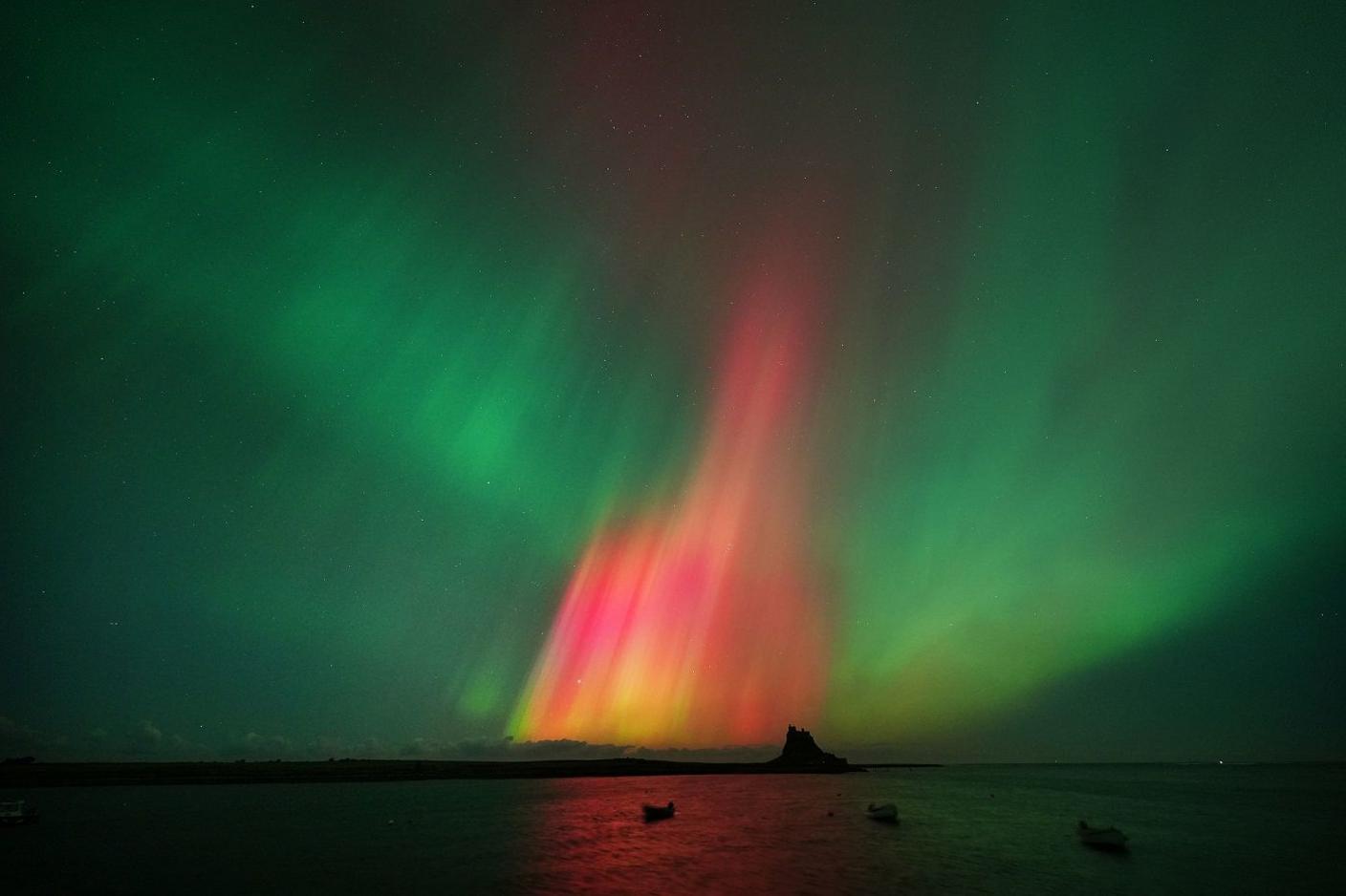 Dramatic red, pink and green light above a silhouette of a castle. Three small boats are on the water can be seen in the foreground.