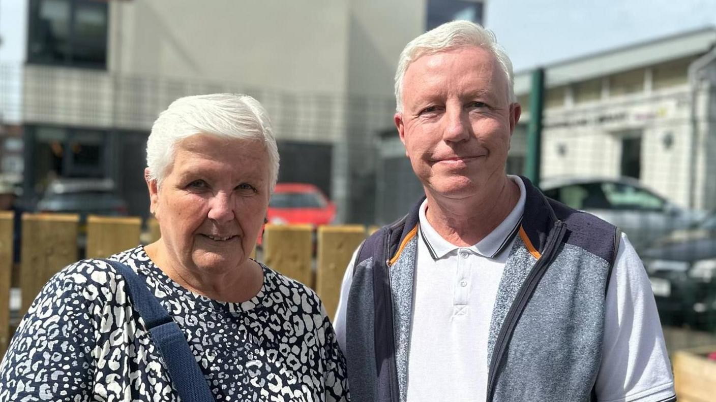 Jean has short grey hair and is wearing a navy and white printed top with a navy bag strap across her body. Next to her is Tom who has short grey hair and is wearing a white polo shirt, grey jumper and is smiling at the camera. Behind them is a brown wooden fence, a metal fence and a number of cars parked.