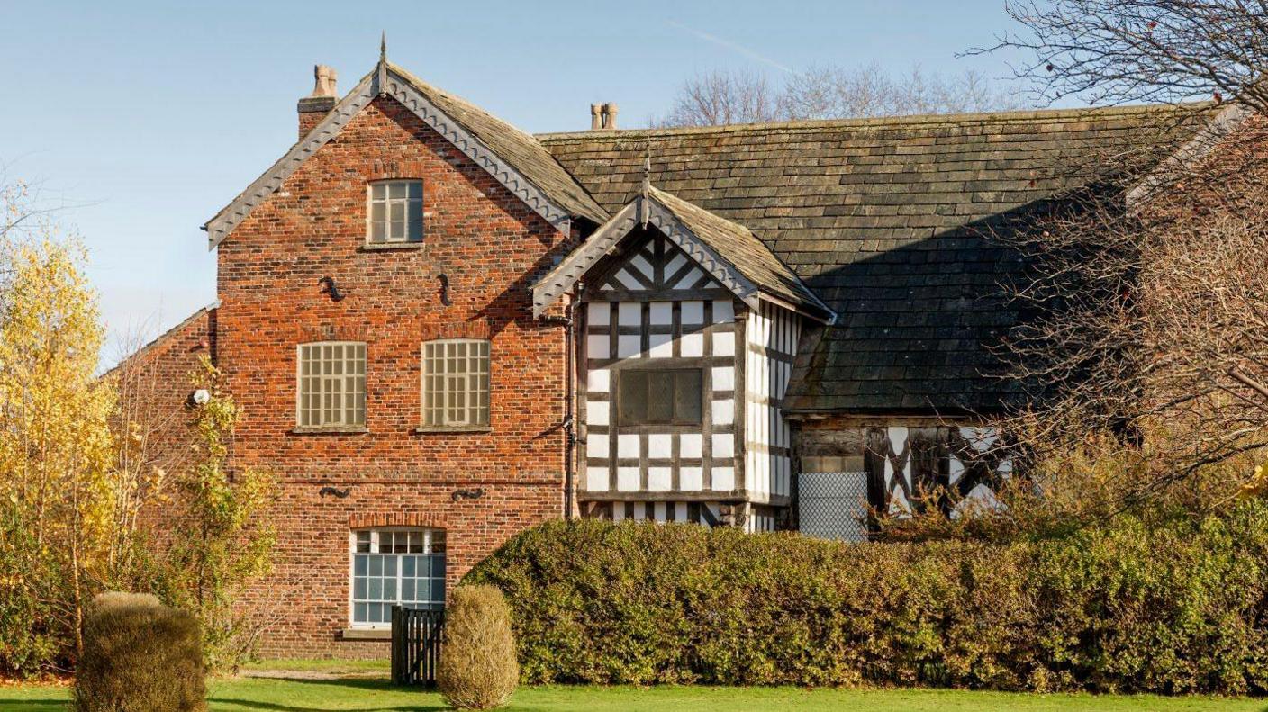 A medieval-style building can been seen, made both of red brick and traditional black and white Tudor architecture. A neat lawn and bushes are visible also