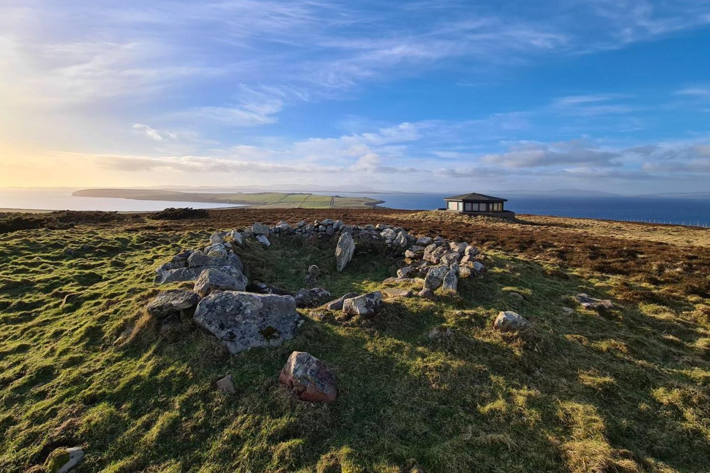 Boulders mark the remains of a Norse beacon in Orkney. There is a modern building to the right and on the horizon an area of low-lying land.