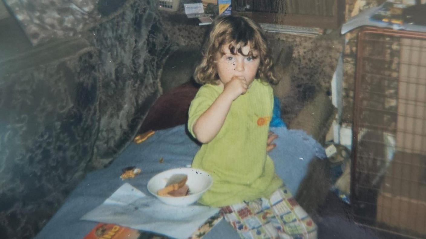 A young girl with curly brown hair, wearing a green top and patterned trousers, is sitting on the sofa. She has her hand up by her mouth.