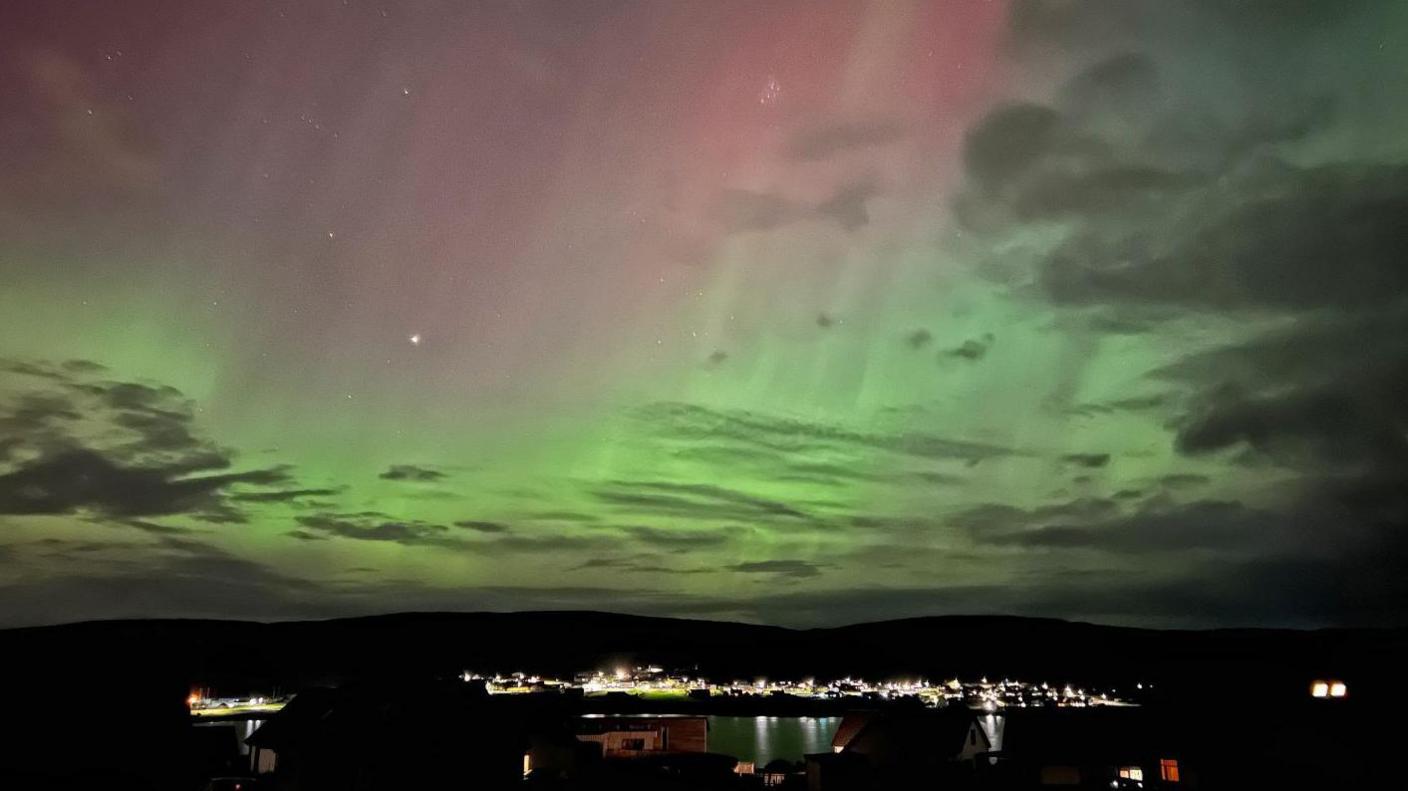 Green and pink aurora shimmering in a cloudy sky above harbour lights.