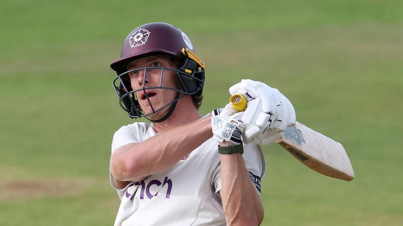 Calvin Harrison of Northamptonshire swings a cricket bat during a County Championship match. He is wearing a white Northamptonshire jersey and a claret helmet with the Northamptonshire rose emblem.