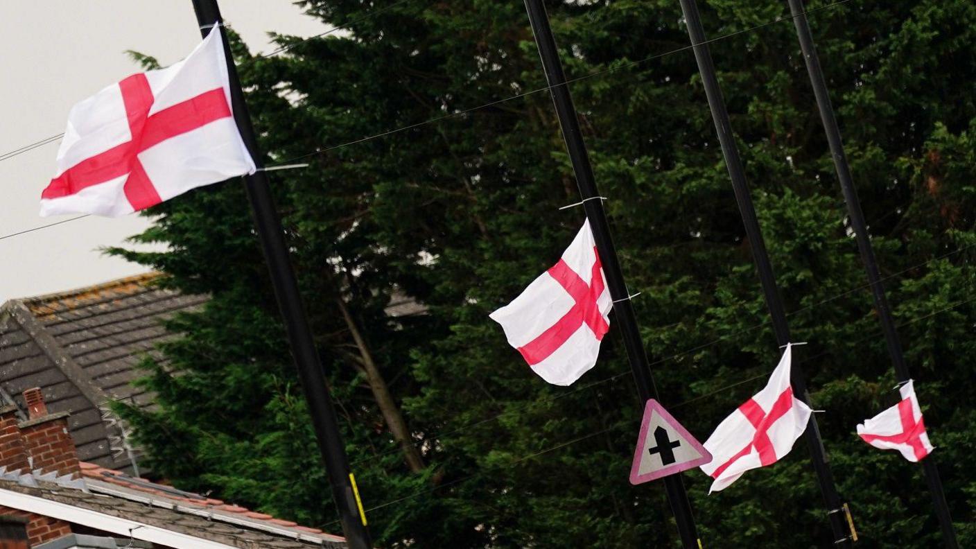 St George's flags fly from a row of black lampposts in Highters Heath in south Birmingham and are rippling in the breeze. Roofs of nearby buildings can be seen to the left and there are trees in the background