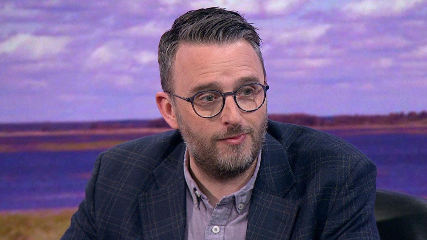 A man with short hair wears circular, black-framed glasses. He is wearing a plaid, dark-coloured suit jacket and a purple collared shirt. He is sitting down in front of a purple-tinged background showing a lake and grass.