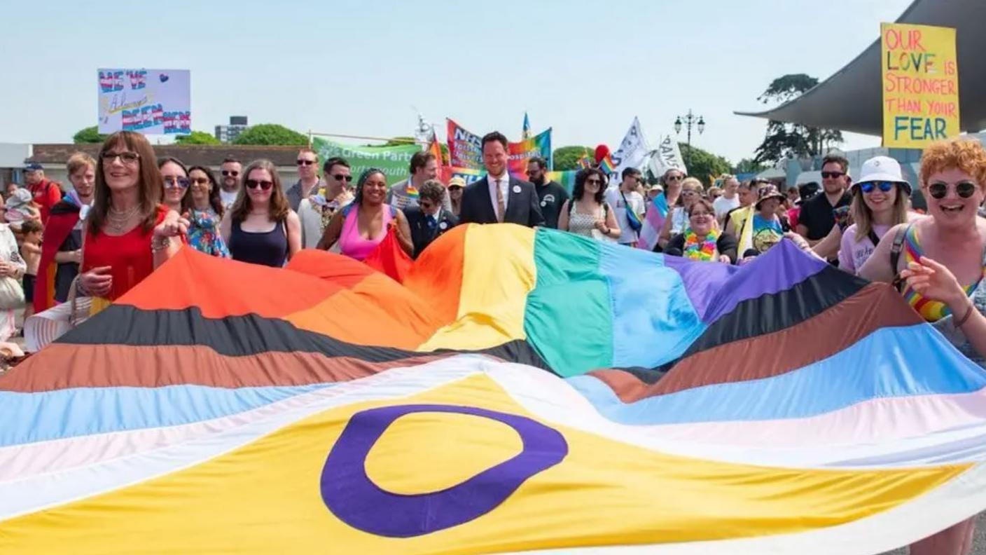 A crowd of people stand behind a Pride flag, which is billowing horizontally, held at the sides by smiling men and women