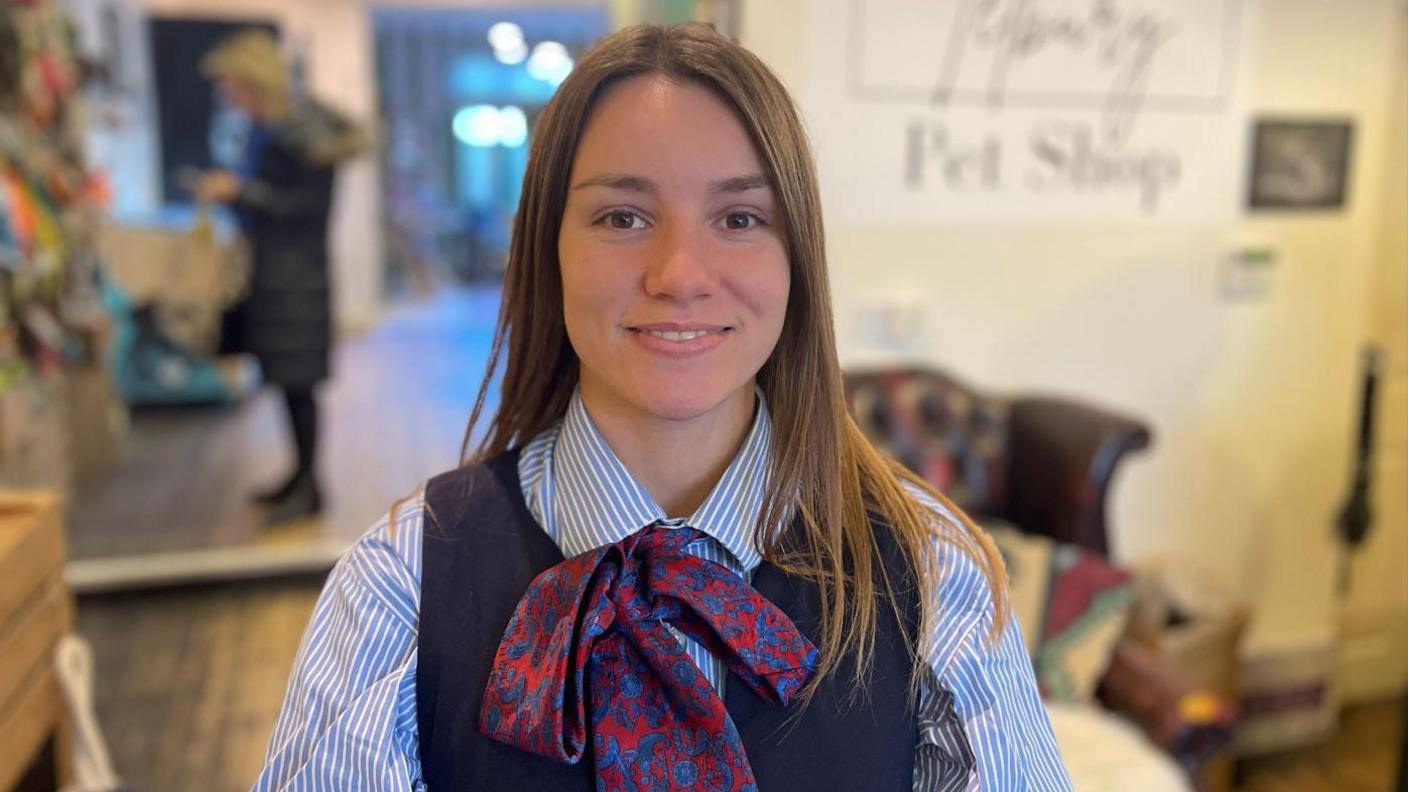 Amelia Black inside the Tetbury Pet Shop. She is a brunette young women wearing a blue striped skirt and a blue pinafore. She is smiling at the camera. 