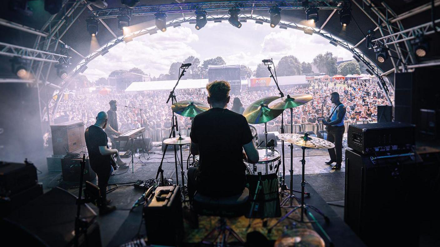 A band playing on a stage, viewed from the back, behind the drummer. In the distance is a crowd,