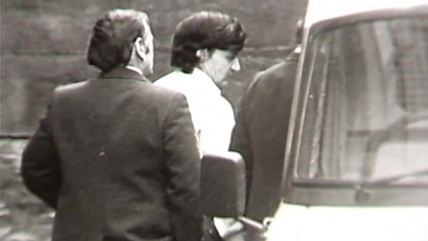 A black and white still from TV footage showing Peter Sullivan, who has thick black hair and is wearing a white shirt, being led to a police van in handcuffs.