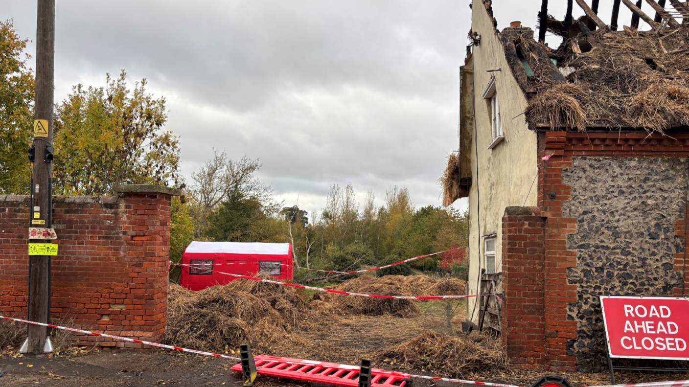 Damage to a thatched roof following a fire. There is tape to cordon off the property and a red "ROAD AHEAD CLOSED" sign is leaning against the cottage.