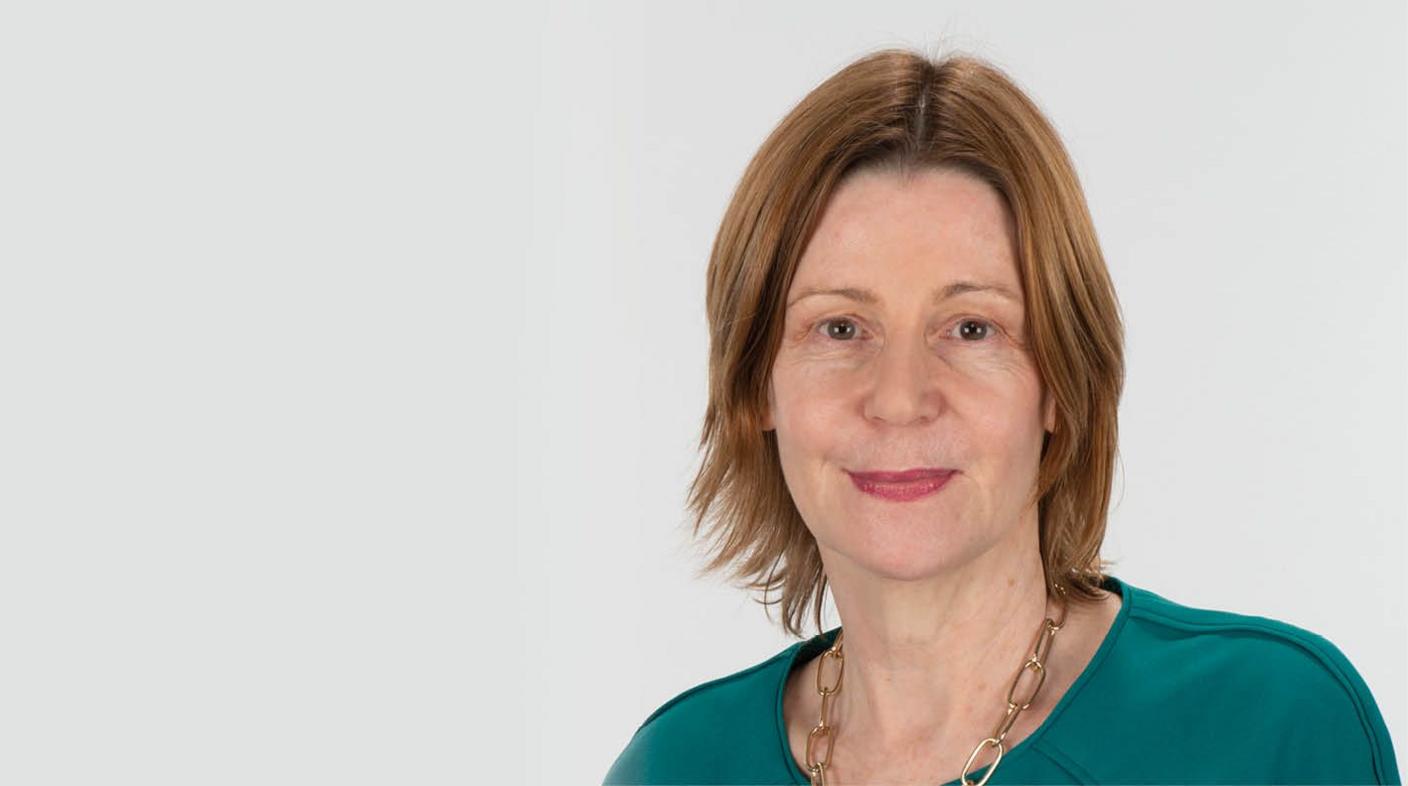 A professional headshot of Maria Kane wearing a teal blouse and a thick gold link chain around her neck. She has shoulder-length strawberry blonde hair and is wearing a dark shade of pink lipstick. She is smiling at the camera and standing against a plain grey background, positioned slightly over to the right.