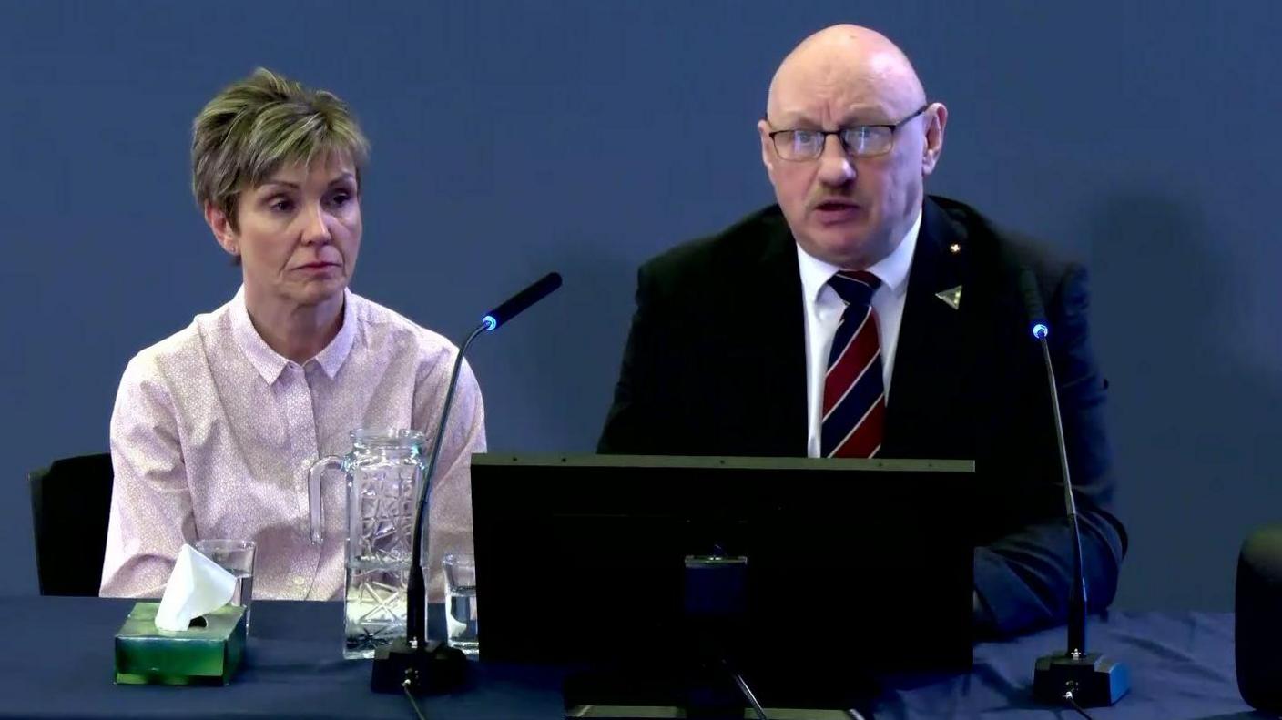 Richard Scott at the Omagh Bombing Inquiry. He sits in front of a laptop and microphones. He wears a blue jacket, white shirt and red and blue striped tie. He is bald and wears glasses. His wife is beside him, with blonde hair. She wears a white shirt.