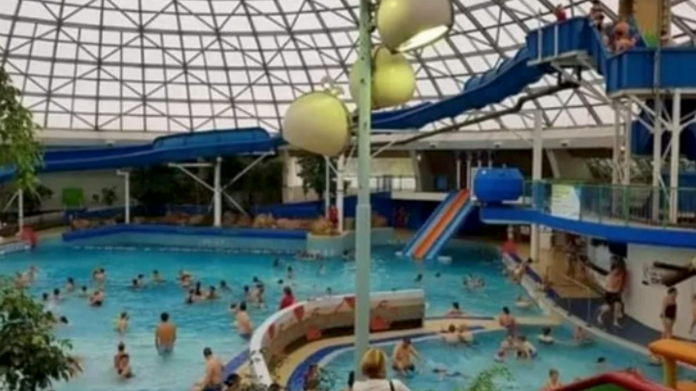 The interior of the Oasis. There is a large blue pool full of people with three big lights. There is also a large blue slide curving down to the left and a smaller slide in the centre.