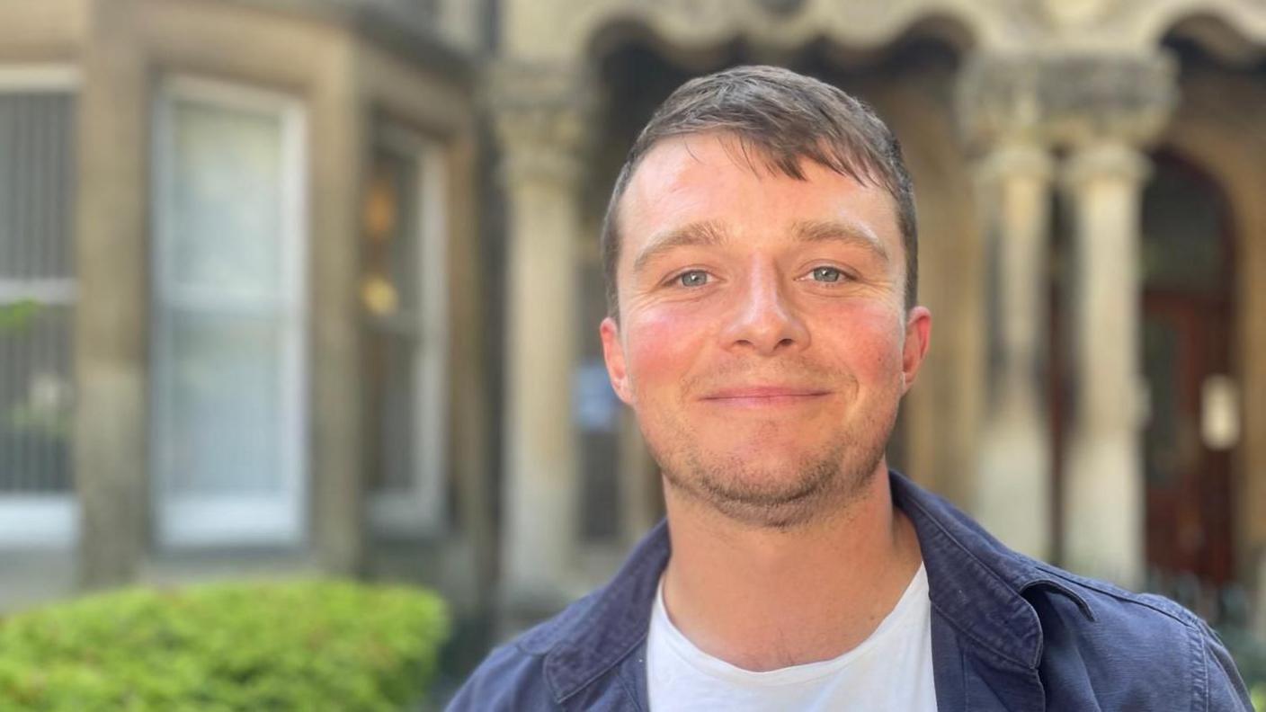 Bill Rowlands, wearing a white T-shirt under a dark blue jacket, smiles outside a large Victorian building.