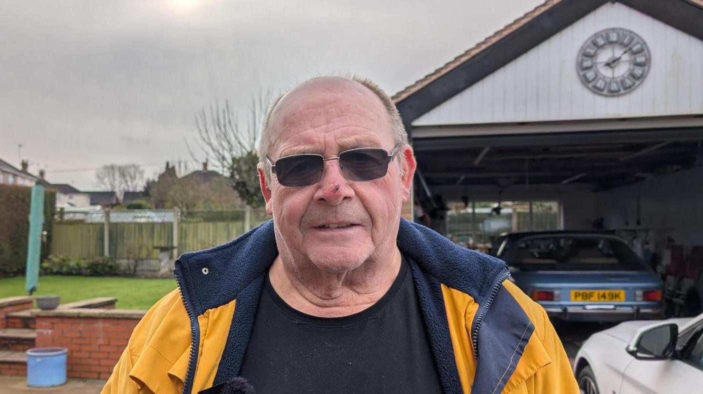 A man in glasses, standing in front of a garage at his home, wears a black t-shirt and yellow coat