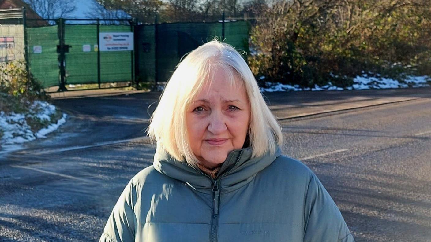 Sue Bussey stands in a blue coat with the entrance to the proposed site behind her