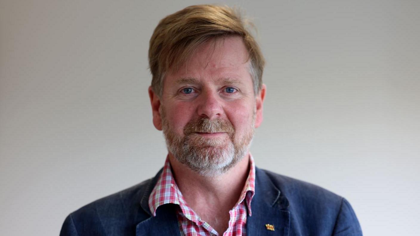 Councillor Nick Oliver, with short brown hair, and a moustache and beard, wears a red and white checked shirt and a navy blazer, with a county council badge on.