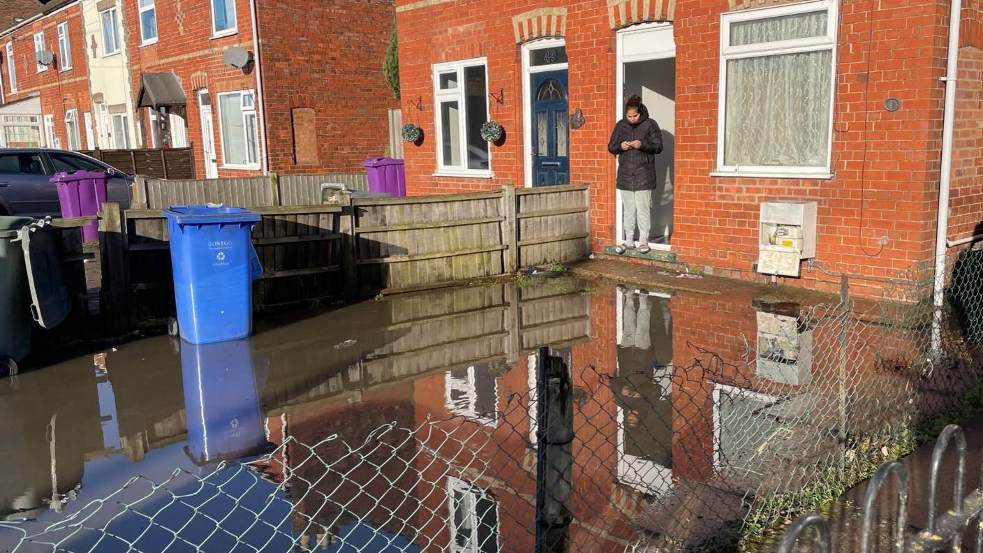 A flooded front garden. At the back is a woman wearing a black coat and grey joggers standing on the front doorstep of a red-brick house looking at her mobile phone, which she is holding with both hands