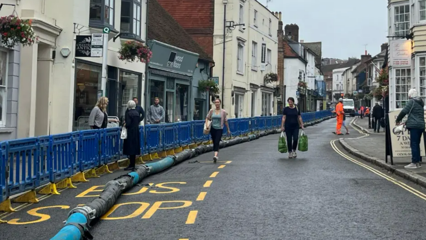 A long pipe snakes down a narrow high street while people walk aside it.