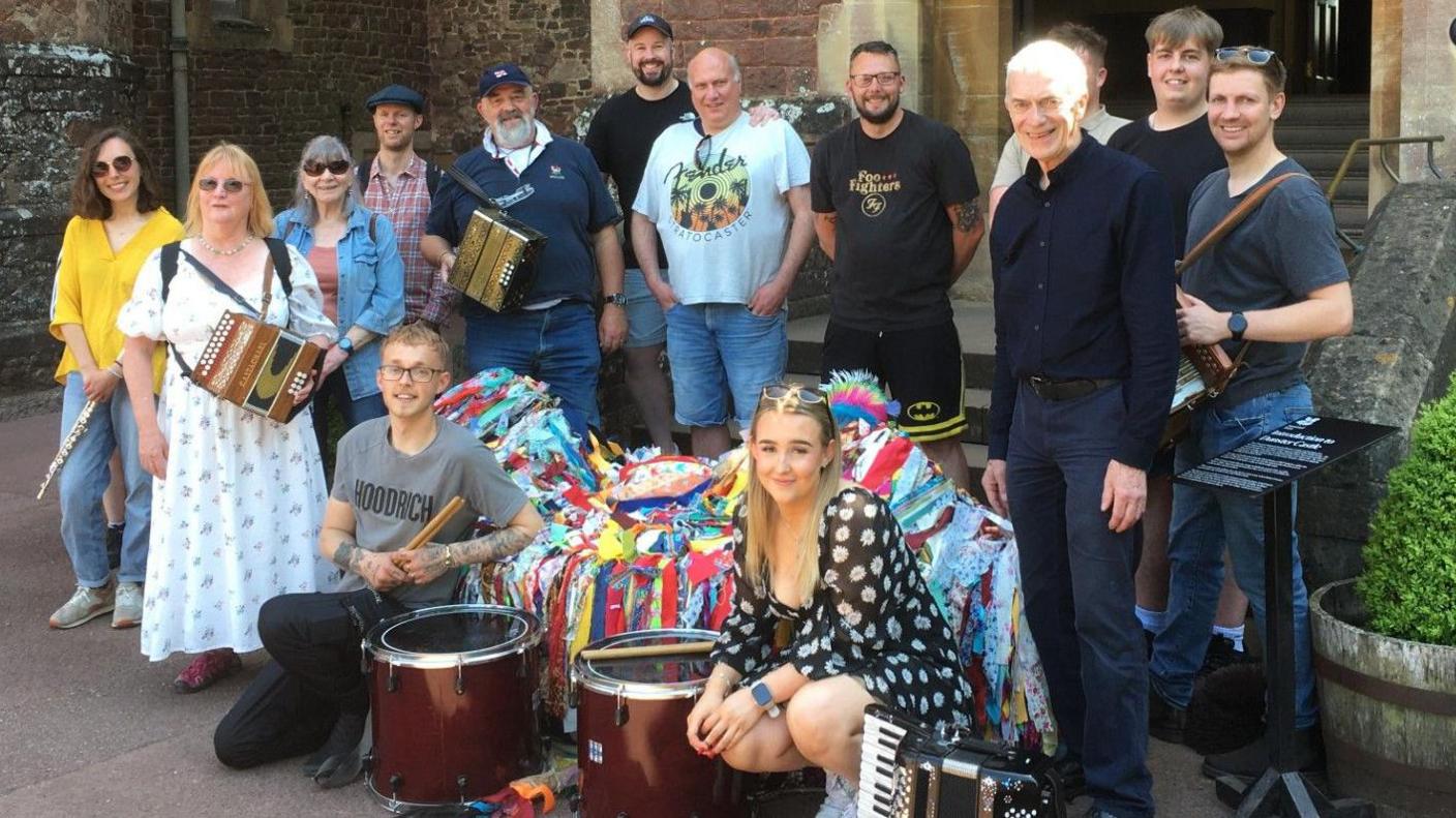 A group of musicians, drummers and followers surrounding a hobby horse. They are of a variety of ages and genders and total about 15 people.